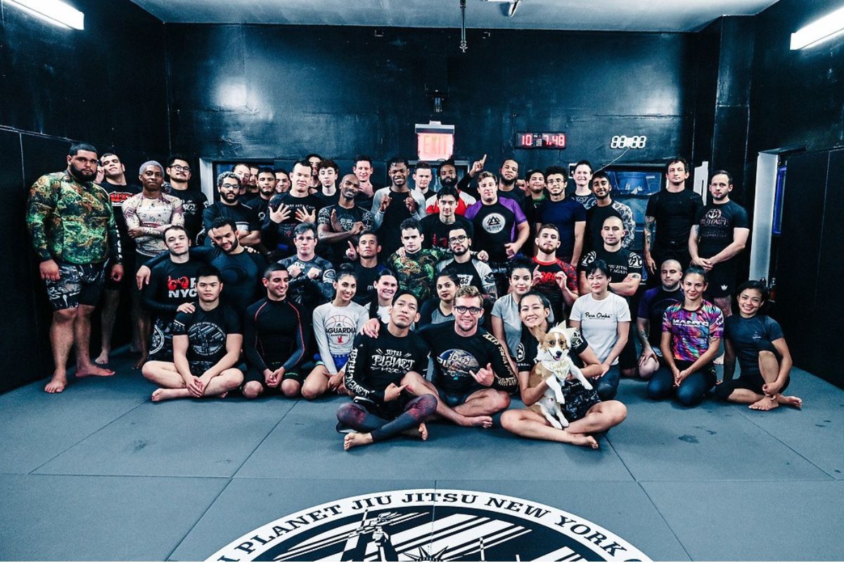 Large group of Brazilian Jiu-Jitsu practitioners posing together in a gym, wearing rash guards and no-gi training attire