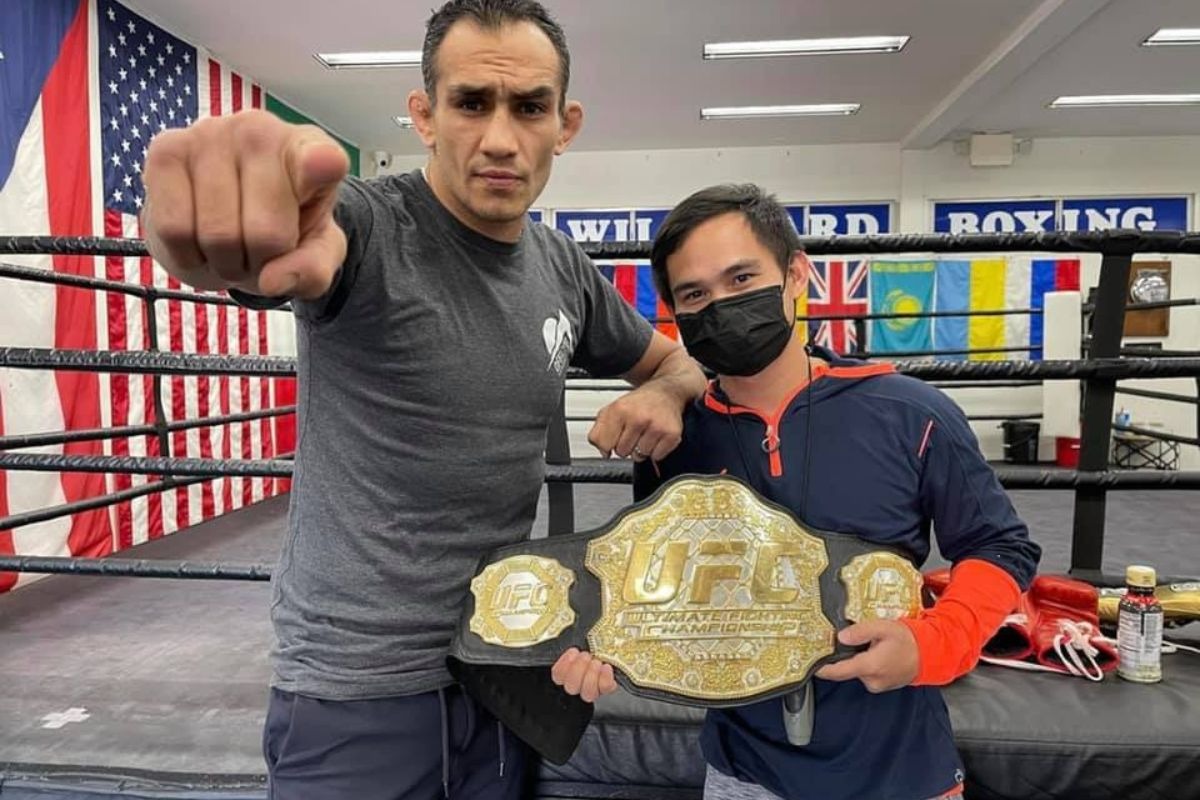 Tony Ferguson poses with a fan holding a UFC championship belt inside a gym, with flags and a boxing ring in the background